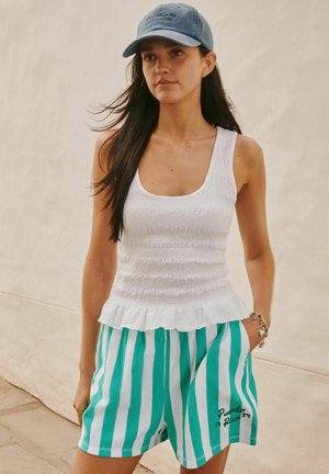 Kobieta z długimi ciemnymi włosami, ubraną w niebieską czapkę, biały bezrękawnik z falbankami oraz turkusowo-białe, pionowo paskowane szorty z napisem "Puerto Rico".