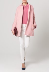 Abrigo de lana rosa claro con un corte relajado, diseño sin cuello y bolsillos laterales, combinado con una blusa rosa y jeans ajustados blancos. Tacones azul marino.
