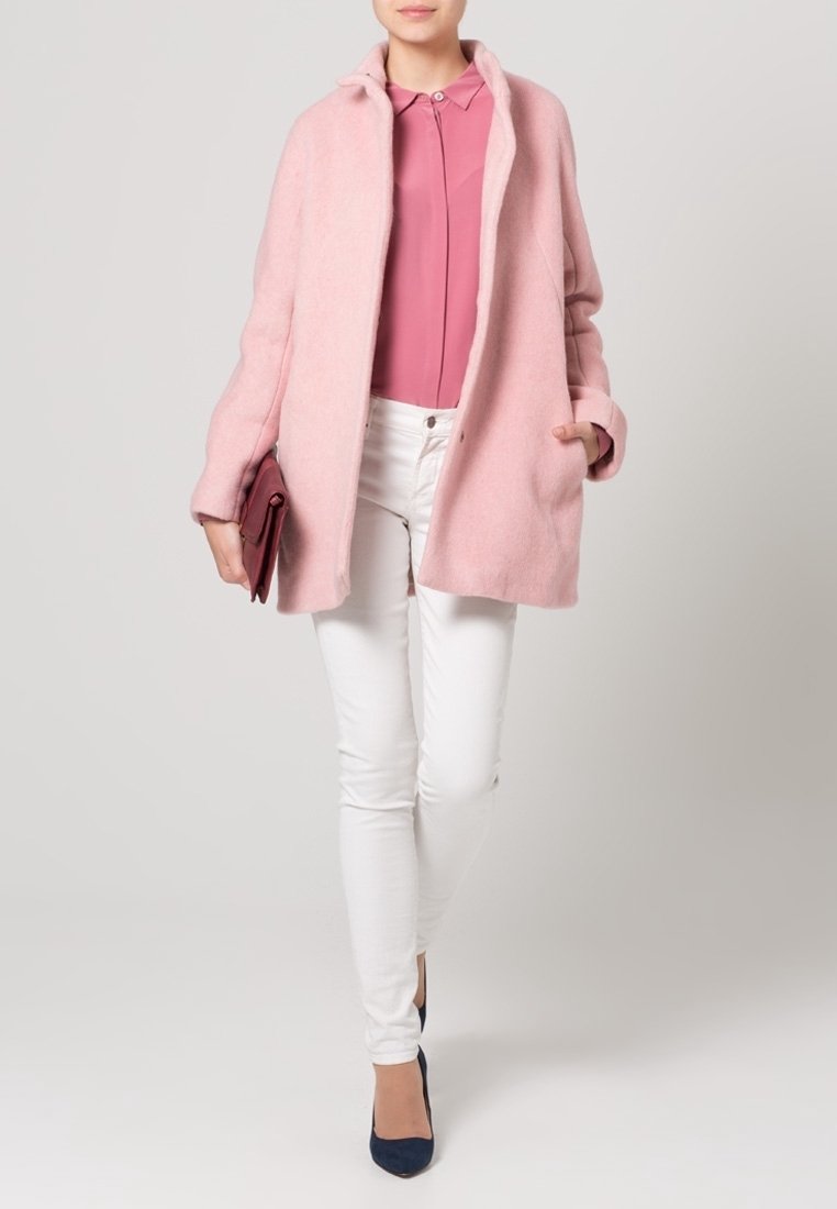 Abrigo de lana rosa claro con un corte relajado, diseño sin cuello y bolsillos laterales, combinado con una blusa rosa y jeans ajustados blancos. Tacones azul marino.