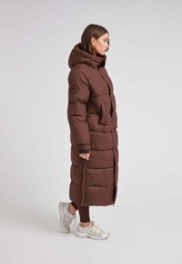 Bruine pufferjas met een capuchon, langwerpig ontwerp en een zijrits. Beschikt over een gestructureerde afwerking en een klein logopatch op de mouw.