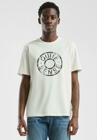 Jonge man in een wit T-shirt met een zwart cirkelvormig "QUIET SENSE" logo, in combinatie met blauwe jeans, staand tegen een effen achtergrond.