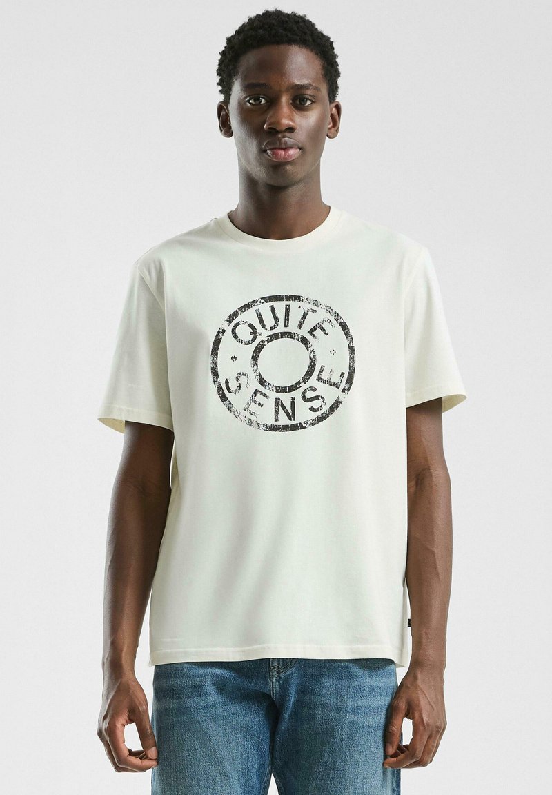 Jonge man in een wit T-shirt met een zwart cirkelvormig "QUIET SENSE" logo, in combinatie met blauwe jeans, staand tegen een effen achtergrond.