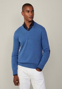 Sininen v-pääntiehen oleva neule pehmeästä neuloskankaasta, ribbaustaskuilla ja helman kanssa, kerroksittain tumman ruudullisen paidan päällä, yhdistettynä valkoisiin housuihin.