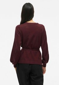 Maglione bordeaux con vita fascia, maniche lunghe e polsini a coste. Presenta una vestibilità comoda e una texture in maglia morbida.