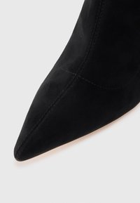 Botine negre din suede, cu vârf ascuțit, având detalii de cusătură subtile de-a lungul laturilor și o textură netedă.