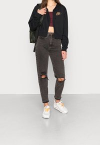 Zwarte hoodie met rits en gouden logo, bordeauxrode crop top, versleten zwarte jeans en witte sneakers met oranje accenten. Groene rugzak op de schouder.