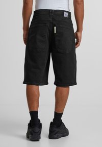 Shorts de mezclilla negra con un corte relajado, que presentan bolsillos traseros y una etiqueta con el logo en la cintura. La tela parece resistente y texturizada.
