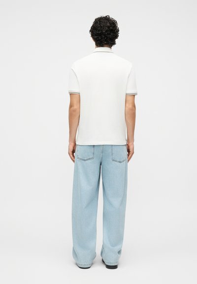 Άνδρας με σγουρά μαλλιά που φοράει λευκό πόλο με κοντά μανίκια και ανοιχτόχρωμο τζιν παντελόνι με φαρδιά πόδια, στέκεται με την πλάτη προς τα εμπρός σε απλό φόντο.