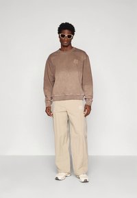 Brown sweatshirt with a graphic design, paired with beige trousers. Model wearing light-colored sunglasses and white sneakers.