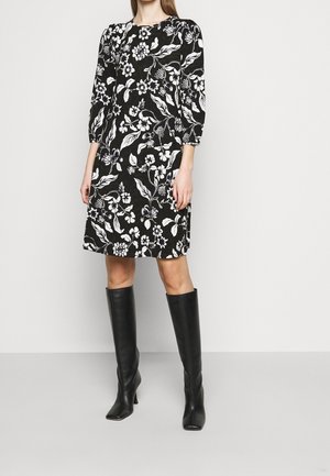 Femme portant une robe noire longueur genou avec des motifs floraux blancs et des bottes noires montant jusqu'au genou à talons, debout devant un fond uni.