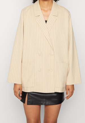 Woman wearing a beige double-breasted pinstripe blazer over a black leather mini skirt with a front slit.