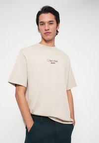 Jauns vīrietis ar tumšiem matiem, ģērbies bēša krāsas Calvin Klein Jeans kreklā un tumšos biksēs, stāv ar rokām kabatās uz baltas fona.