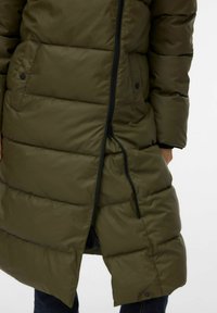 Olivirohelisest polsterdatud jope lukuga, millel on horisontaalsed viltseotud sektsioonid, küljetaskud ja lõhestatud ääreosa liikumiseks.