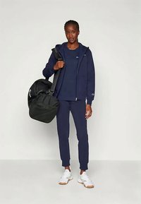 Navy Zip-Jacke, navy T-Shirt und passende Jogginghosen, kombiniert mit weißen Sneakers. Hält eine schwarze Sporttasche mit Logo-Detail.