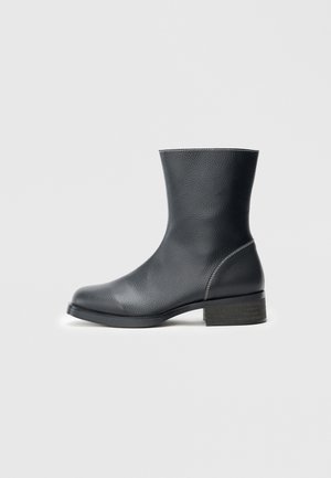 Botín negro de cuero con acabado texturizado, punta redonda y un tacón bajo de madera. Costuras claras acentúan los bordes.