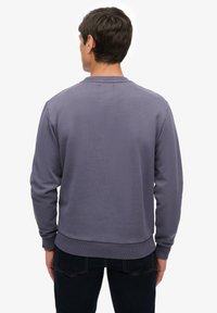 Fioletowa bluza z teksturowanym wykończeniem, prążkowanym kołnierzem i mankietami. Dopasowany fason z prostym dołem, zestawiona z ciemnymi jeansami. Widok od tyłu.