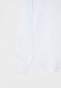 Camicia bianca a maniche lunghe con una texture liscia, polsini sartoriali e un orlo leggermente curvato. Design semplice e minimalista.