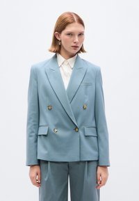 Ανοιχτό μπλε διπλό σακάκι με δύο τσέπες μπροστά, κουμπιά σε αντίθεση και πέτο με σχισμή, συνδυασμένο με παντελόνι στο ίδιο χρώμα.