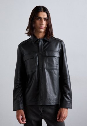 Young person with long dark hair wearing a black leather button-up jacket with two chest pockets and dark pants, standing against a gray wall.