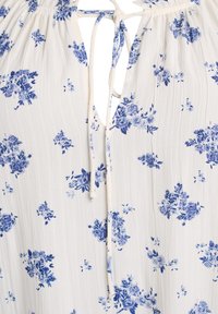 Blusa floreale in tessuto crema con motivi floreali blu. Presenta una chiusura con laccio e una superficie testurizzata. Vestibilità ampia e design leggero.