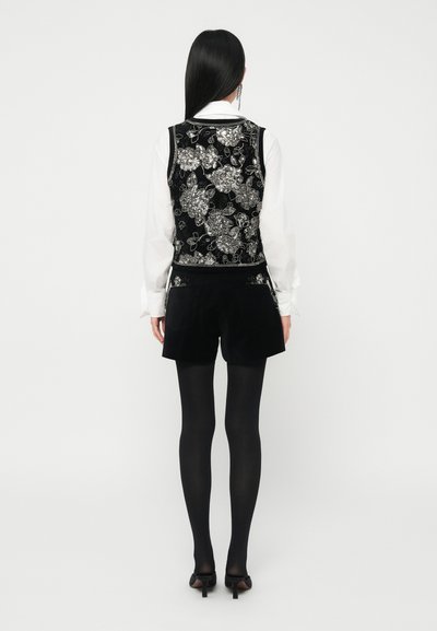 Femeie cu părul lung și negru stând cu spatele, purtând o bluză albă, o vestă brodată floral neagră, pantaloni scurți negri, colanți negri și pantofi cu toc negri.