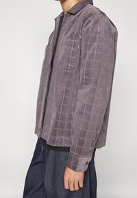 Chaqueta de pana en color púrpura con un patrón de cuadros, que cuenta con un cierre en la parte delantera con cremallera, dos bolsillos en el pecho y puños abotonados.