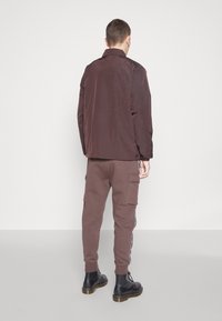 Hnědá lehká bunda s límečkem, kombinovaná s hnědými cargo kalhotami s bočními kapsami a pruhovanými detaily. Černé šněrovací boty.