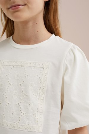Close-up af en person iført en hvid top med puffede ærmer og et broderet firkantet blondemønster på brystet.