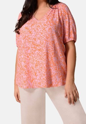 Kvinde iført en lyserød V-hals bluse med orange blomsterprint og beige bukser, stående med den ene hånd hvilende på sit lår.