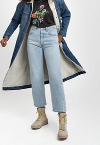 Denimjacka med shearling-fodring, svart grafisk T-shirt, ljusblå raka jeans, beige vattentäta kängor med gula snören.