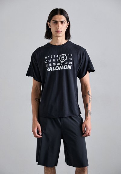 Černé tričko s krátkým rukávem s kosticovým vzorem, se stříbrným číselným designem a textem "SALOMON". V kombinaci s odpovídajícími černými šortkami.