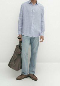 Man in een lichtblauwe gestreepte overhemd met knopen en jeans, die een grote donkerbruine suède tote bag vasthoudt, staand op een witte vloer.