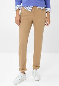 Beige Chinos mit Slim Fit, die eine glatte Textur, Seitentaschen und hochgekrempelte Säume aufweisen. Kombiniert mit weißen Sneakers und einem blauen Pullover.