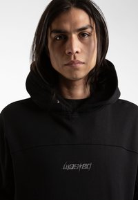 Black hoodie with a high neck and drawstring. Features embroidered "wasted" logo on the chest, textured fabric, and a relaxed fit.