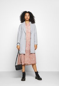 Lichtgrijze jas met een reverskraag, gedragen over een roze blo dress. Zwarte legerboots en een kleine zwarte schoudertas maken de outfit compleet.