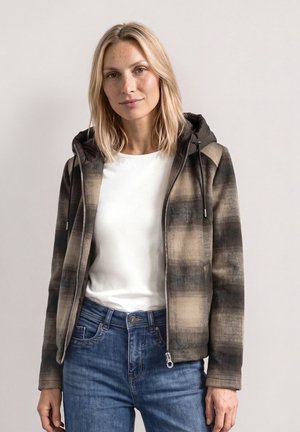 Femme blonde portant une veste zippée à carreaux marron sur une chemise blanche et un jean bleu, debout devant un fond clair uni.