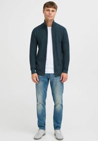 Dark teal zip-up jacket with a stand collar, worn over a white shirt; paired with light blue jeans and gray sneakers. Casual fit and texture visible.
