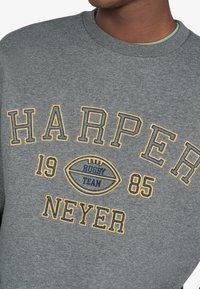 Sudadera gris con bordado amarillo y azul marino que dice “HARPER NEYER 1985 RUGBY TEAM” y un gráfico de un balón de rugby.