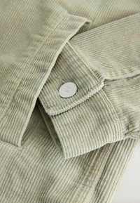 Tissu en velours côtelé beige clair à nervures verticales, présentant un revers plié maintenu par un bouton argenté orné d'un petit logo gravé.