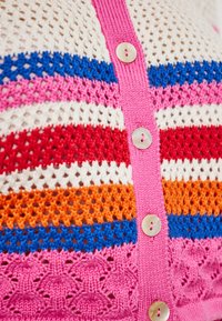 Megzta kardiganas su horizontaliomis juostelėmis: rožinė, raudona, oranžinė, mėlyna ir balta. Keturi apvalūs mygtukai su tekstūruota raštine ant audinio.