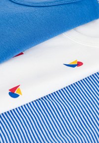 Trois t-shirts en coton empilés : un bleu uni, un blanc avec un logo de voile multicolore, et un rayé bleu et blanc.