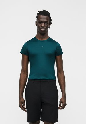 Tummaihoinen, letitettyä tukkaa oleva mies pukeutuneena vartalonmyötäiseen teal-vihreään lyhythihaiseen paitaan ja mustiin shortseihin, seisoo yksinkertaisen vaalean taustan edessä.