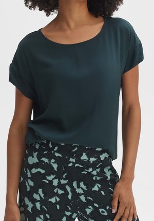 Mørkegrøn kortærmet bluse med rund halsudskæring; glat tekstur. Sammen med sorte og mintfarvede mønstrede bukser med blomsterprint.