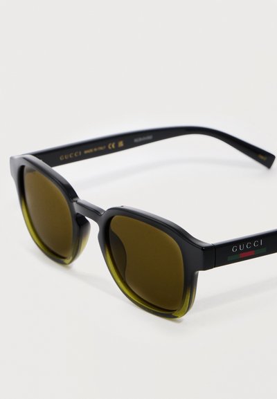 Lunettes de soleil Gucci noires avec verres teintés carrés et logo vert-rouge-vert sur la branche, posées sur une surface blanche.