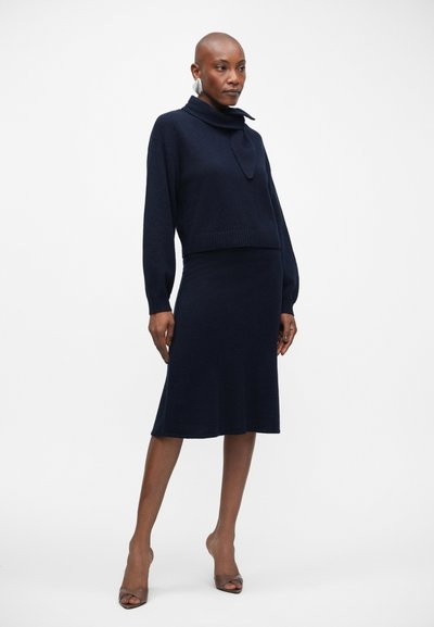 Conjunto de suéter e vestido em azul-marinho, feito de um tecido suave e texturado. O suéter possui uma gola alta e bainha canelada; o vestido é ajustado e de comprimento até ao joelho.