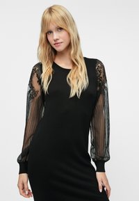 Juoda megzta suknelė su permatomomis juodomis nėrinių pūstomis rankovėmis, kurių sudėtyje yra gėlių raštų ir rausvų rankogalių, sukuriančių tekstūrų kontrastą.