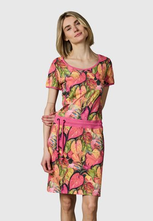 Frau in einem bunten, kurzärmeligen Kleid mit Blättermuster, rosa Taillenband und Kordelzug, stehend vor einem schlichten hellen Hintergrund.