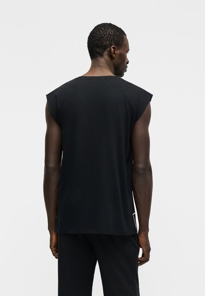 Άνδρας με σκουρόχρωμο δέρμα, ντυμένος με μαύρο αμάνικο πουκάμισο και μαύρο παντελόνι, με την πλάτη γυρισμένη, σε απλό ανοιχτό γκρι φόντο.