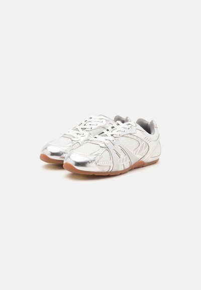 Paire de chaussures de sport blanches et argentées avec des lacets blancs et des semelles marron clair sur un fond uni.