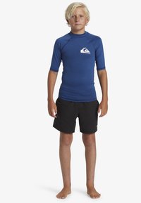 Blå kortärmad rash guard med en logotyp på bröstet, i kombination med svarta badshorts. Slät textur, åtsittande design och inga mönster.
